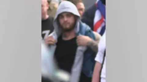 Northumbria Police A blurred image of a man in a hooded top, with a brown beard and dark hair, who is wanted by police. He is wearing a black top and grey hoodie, and has a sleeve tattoo down his left arm.