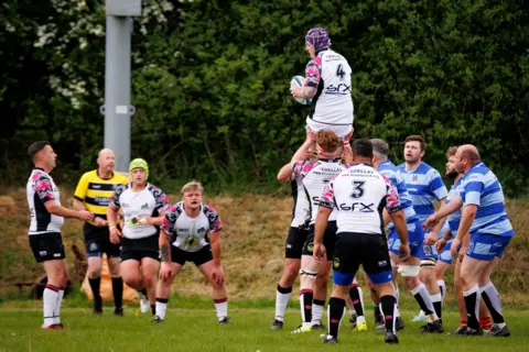 Handout A match between the Hospice Barbarians and The Gorillas RFC. Players from both teams are seen in their different kits. One man is on another man's shoulders. They are on a rugby pitch with a post and trees in the background.
