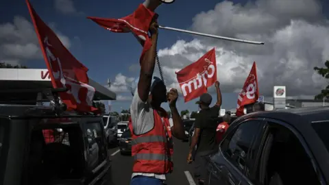 PHILIPPE LOPEZ/AFP Men in gilets wave flags of the French union CGT Martinique during aa go slow operation on the ring road leading to the airport in Fort-de-France, in the French Caribbean island of Martinique.