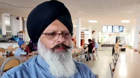 Harjap Sing, a man with a long white beard and black moustache, is wearing a blue collared shirt and a black turban. He is sat at a long table in a room that resembles a town hall or community centre. Other people are sat at tables around the room. 