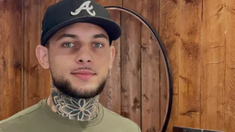 Stephen Huntley/BBC A man with a short beard wearing a baseball cap looks directly at the camera. He is standing in a barber shop and has a distinctive flower-based neck tattoo.