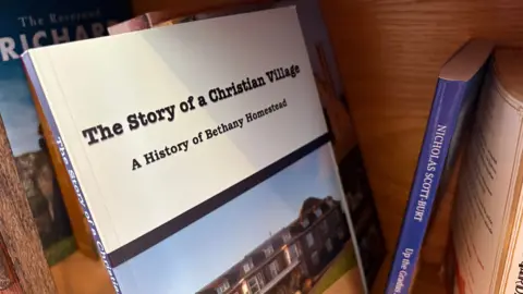 Martin Heath/BBC Book with a beige cover entitled "The story of a Christian Village - a History of Bethany Homestead". It has a picture of a three-storey building on the site. It is pictured on a bookshelf with other books.