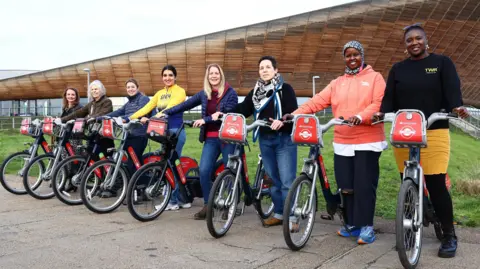 Eight women each holding a Santander TFL hire bike, which has been named after them for being an inspirational women in the cycling community. The women are all smiling