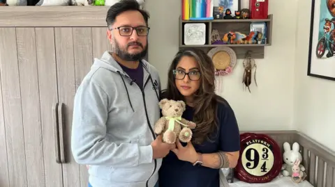 A man and woman stand together in a child's nursery holding a teddy bear. The woman has long dark hair and glasses. The man also has dark hair and is wearing glasses. 