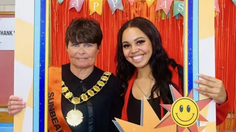 Klarisse Smith Klarisse and the Lord Mayor of Nottingham smiling and posing with a decorative photo frame in front of a red backdrop with colourful hanging ribbons and bunting. The Lord Mayor of Nottingham, on the left, is wearing a gold civic chain, a black outfit, and an orange sash reading "SUNBEAM". Klarisse, on the right, has long dark hair and is wearing a red blazer over a black top. A large cut-out sun with a smiley face is attached to the photo frame.