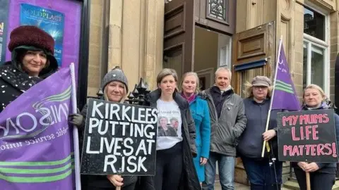 Seven people stand outside Huddersfield Town Hall holding placards saying 'Kirklees putting lives at risk' and 'My mum's life matters'.