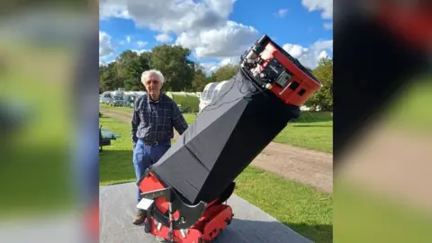 Alan Marriot, wearing a check shirt and blue trousers. He is standing next to a large red and black telescope which is taller than he is. 