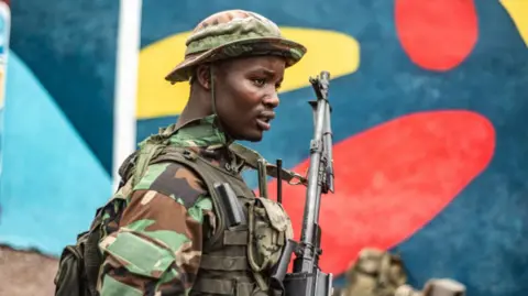 A soldier wearing army fatigues and carrying a gun stands in front of a colourfully-painted wall.