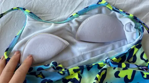 Multi-colour swimsuit with neon yellow and black leopard print hearts on a blue base colour. The swimming costume is laid flat and the inside layer is shown from behind. The inside is white and over the chest area there are oval-shaped padded cut-outs.