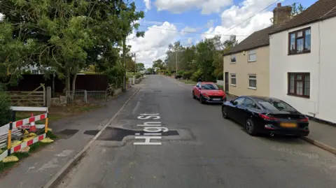 Google Street View High Street in Cherry Willingham showing several tarmac patches where repairs have been carried out. There is a traffic cone and temporary fencing on the left hand side of the pavement. 