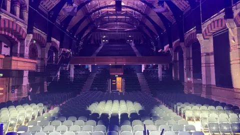 Interior of the Cambridge Corn Exchange. It has seats in rows, as well as on a balcony and brick walls to the side.