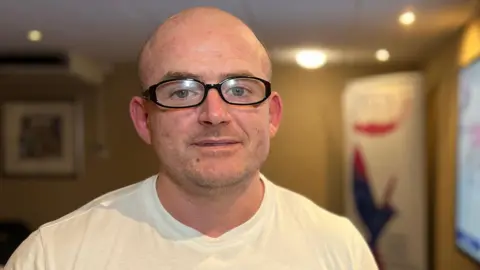 Kevin is wearing a white T‑shirt with a name tag reading “Kevin” stands in a meeting room. A large screen displaying colorful word art is visible in the background, along with a coffee cup, laptop, and presentation materials on a table.