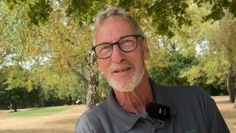 BBC/Lilah Parsons Jim Boise wearing glasses and a grey polo shirt with a small logo, standing beneath leafy trees beside a golf course, with dry grass and mature trees visible in the background.