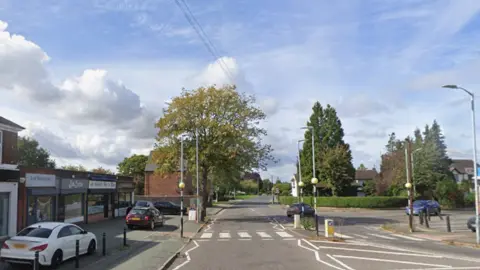 Google Maps General view of Langley Road in Lower Penn, Wolverhampton. A zebra crossing is in the middle of the picture and on the left-hand side there is a row of shops and parked cars. In the background there are trees and hedges as the road continues