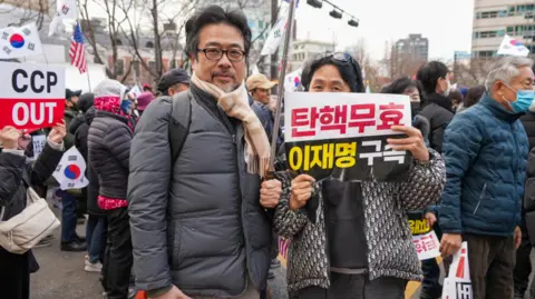 BBC/Hosu Lee A man stands with a woman, who holds a sign saying that the impeachment was invalid