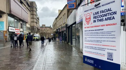 Close-up of a poster on a lamppost detailing the use of live facial recognition technology. In the background people walk past along a wet street lined with shops. A live recognition van can bee seen in the distance. 