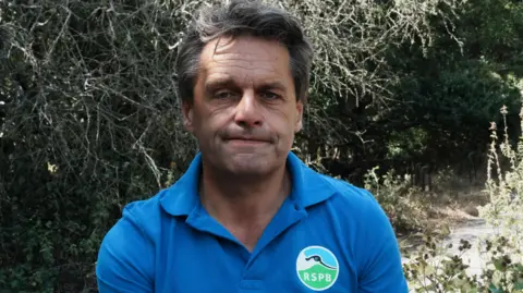 John Fairhall/BBC A head-and-shoulders image of a man standing in front of burnt hedging. He has a look of resignation. He is looking straight at the camera. 