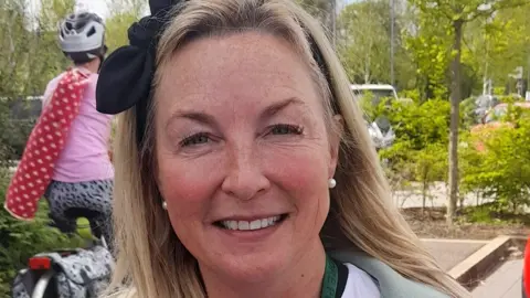 Wendy Lyons has long blond hair with a black ribbon in it and is smiling. A cyclist is in the background of the picture.