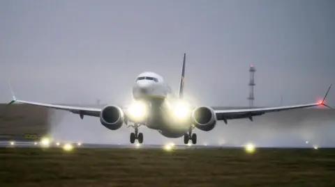 Danny Lawson/PA Wire Plane Landing di Bandara Leeds Bradford, dengan lampu pendaratan bersinar ke kamera dan banyak air hujan di belakangnya, sementara menara transmisi berdiri di latar belakang.