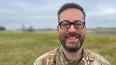 Amber Macey/BBC Wing Cdr Colin Melvin wears a camouflaged outfit and rectangular black glasses. He is standing b a green field with a plane in the background and  smiling into the camera