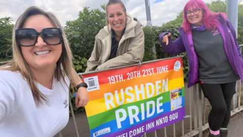 Rushden Pride Three women, all leaning on a Pride banner, by railings. They are all smiling and looking at the camera. The woman on the left has sunglasses on and a white T-shirt, the woman in the middle, long hair, tied up and wearing a cream jacket, and the woman to the right has pink hair, is wearing a purple jacket, black top and black leggings. Bushes are behind them. 