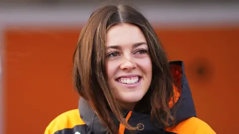 Getty Images Ella Stevens smiling while wearing a black and orange raincoat.