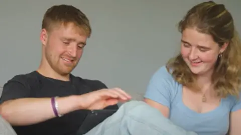 Children with Cancer UK Two siblings sitting next to each other on a sofa. Male, Seb in a navy t-shirt and jeans and female, Libby in a pale blue t-shirt both smiling at each other.