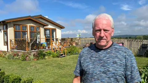 A white man with grey hair. He is wearing a blue top. It is a sunny day and in the background is his mobile park home.