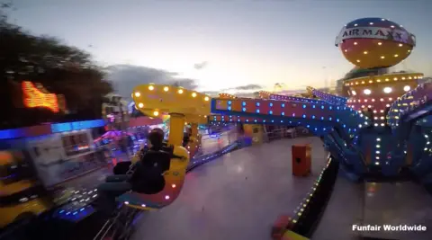 The Air Maxx is captured in motion during dusk. The ride consists of large mechanical arms decorated with vivid lights in blue, yellow, and pink. One of the arms is elevated, holding a seat with two people securely strapped in, facing outward. The background reveals additional illuminated rides and attractions, contributing to the festive atmosphere. On the left side, tall trees frame the scene, while the sky above is partly cloudy, transitioning from daylight to evening.