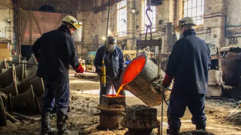 Loughborough Bellfoundry Trust Three men stand pouring molten liquid into a hold. They are in the workshop and wearing blue overalls. 