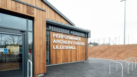 Sport England The outside entrance of a modern building, clad in brown wood, with Performance Archery Centre Lilleshall written on it in white lettering. Bike racks can be seen to the right and the path is tarmacked. A bare earth slope leads up to sports courts.