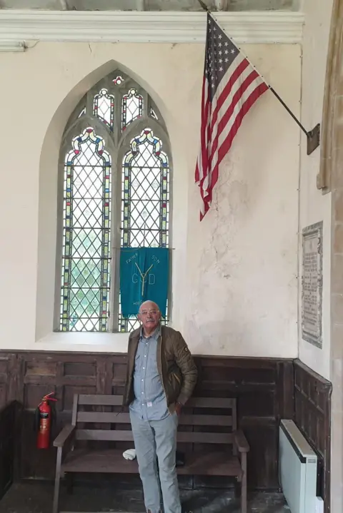 John Stockley standing in front of a church window with the US stars and stripes flag attached to the wall above 