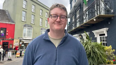 BBC News Frankie Bennet, wearing glasses and a blue quarter zip jumper and a light green t-shirt.