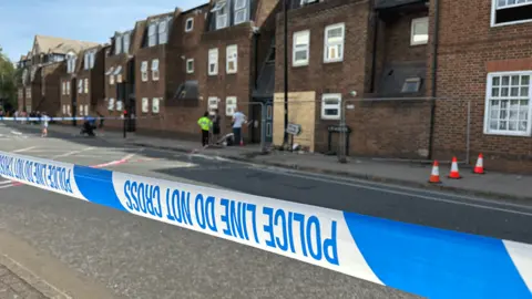 A view of the same scene, further back, from the vantage point of the blue and white police cordon.