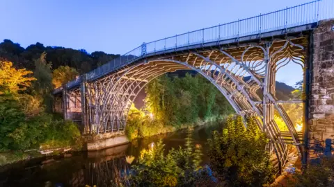 Getty Images An elaborate metal-framed bridge high over a river with trees on the far side, illuminated by lights shining up at it