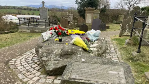 A large boulder in the grounds of Down Cathedral is covered with floral tributes as the stone marks the reported burial place of St Patrick. It is surrounded by cobbled paving and is marked with a stone plaque.  Other headstones are in the graveyard and the Mourne Mountains can be seen in the distance. 