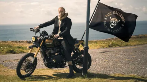 Sam Heughan in a black jacket sits on a motorbike in front of the sea. The bike has a flag saying "RIDE GALLOWAY DARE TO EXPLORE"