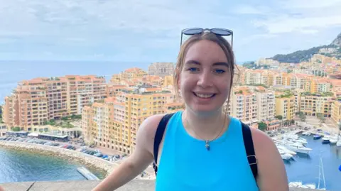 Claire Thomson Claire standing in front of high-rise buildings in Monaco. She is wearing a blue top with her hair tied back and black sunglasses on her head.