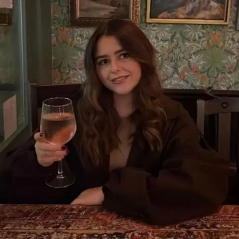 Analise Taylor A woman with long, wavy, chestnut hair smiles at the camera and holds a glass of wine.