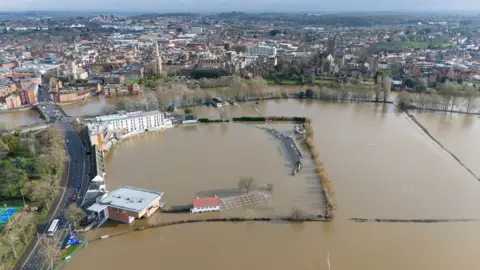 PA Media An aerial view shows the cricket ground is pictured under brown water, with rugby posts popping up above the water line next to it and the backdrop of the town of Worcester, taken on Monday.