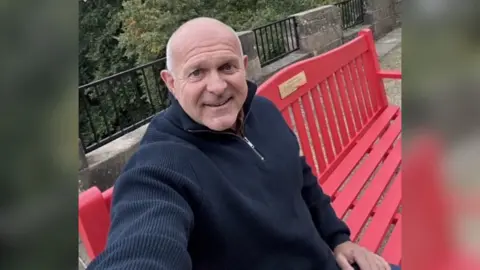 Dean Dyble Dean Dyble sits on a bright red wooden red bench. He is an older man, with a bald head and a blue jumper, blue jeans and brown boots. In the background, trees and a stone wall can be seen.