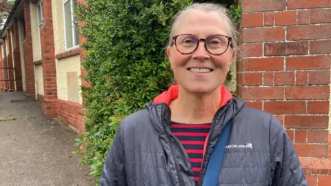 BBC/Carla Fowler Mullane is standing close to the camera, facing forward. She is wearing a dark quilted jacket with a visible brand name printed on the chest that reads “ENDURA”. Underneath the jacket, a striped top with red and dark horizontal stripes can be seen. A crossbody strap runs diagonally across her torso.
She is also wearing round glasses with dark frames, and her hair is tied back.