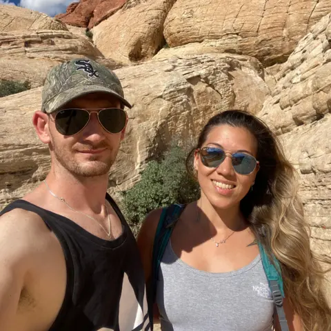 Kelly Burston A selfie of a couple - a man and a woman - taken in front of large rocks outdoors. The woman is wearing a grey top and is smiling at the camera. The man is wearing a black tank top and a cap. Both are wearing sunglasses