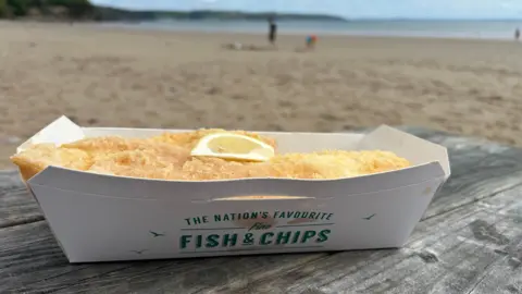 Cod, with a slice of lemon, in fish and chips tray on bench looking out at the sea