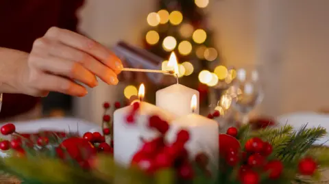 A person lights a white candle with a match on a festive table decorated with holly and red berries, with a softly lit Christmas tree blurred in the background.