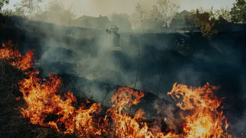 Edward Matthews Flames rip through a crater. A firefighter can be seen through the smoke.