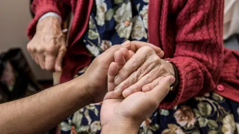 Getty Images A pair of hands holding the hands of an elderly woman. The woman is wearing a red cardigan and a dress with a floral print. She is sitting.