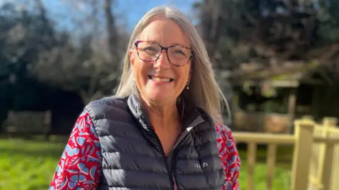 A woman with long grey hair is standing on decking in the sun outside the MS centre. She is wearing glasses along with a dark blue gilet on top of a flowery blouse. She is smiling. 