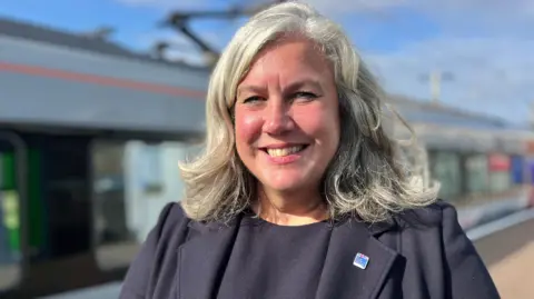 Heidi Alexander smiles at the camera, she is standing on a train platform in the sun, with the train behind her.