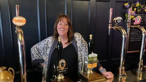 Hazel Smith stands behind the bar set up in a tap room created in the couple's front room. There are tap handles customised with brewery branding and there is a board with writing on to one side, decorated with flowers. Mrs Smith is wearing a black top and a patterned cardigan. She is smiling, with one hand on the bar, and has shoulder-length hair.